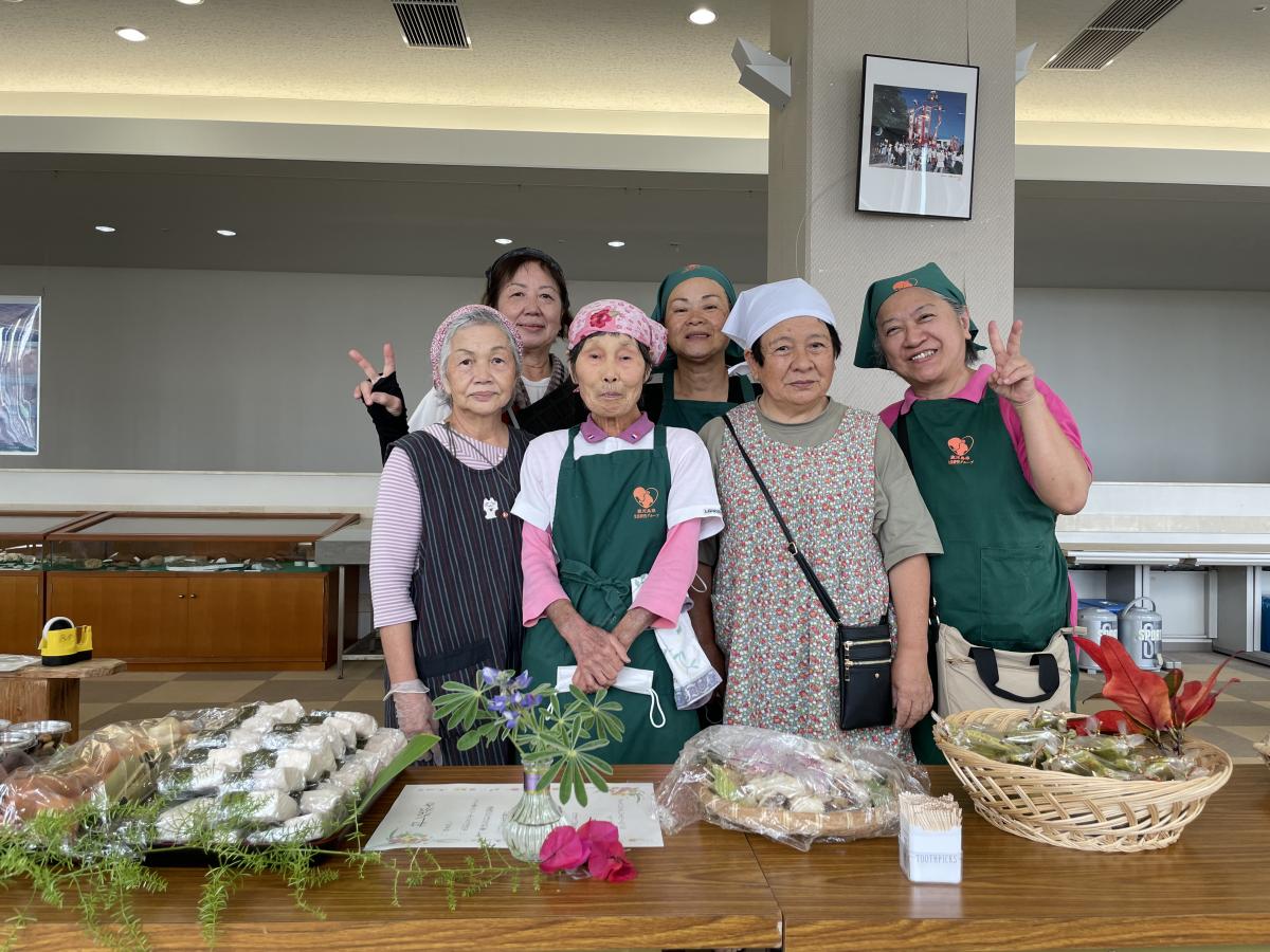 緑のエプロンと様々な服装をした6人の女性たちが、笑顔でピースサインをし記念撮影している写真