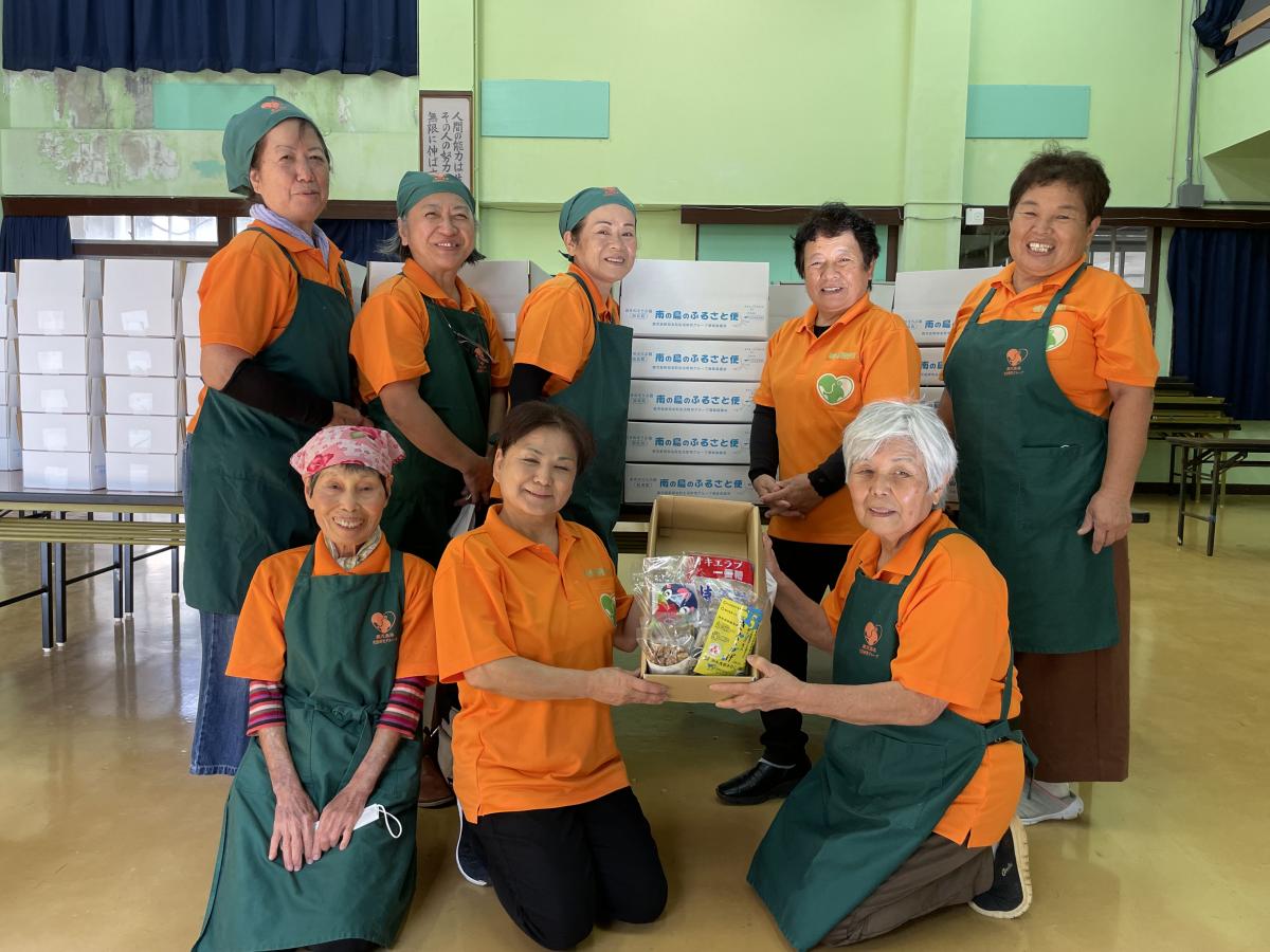 室内でオレンジ色のポロシャツと緑色のエプロンを着用した参加者達が集まり、中央2人が箱に入った食料品を持ち記念撮影している写真