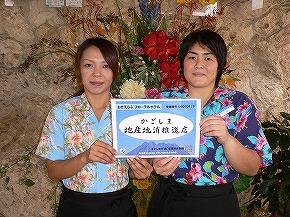 花を背景に、色鮮やかなアロハシャツを着た二人が並び、中央で「かごしま地産地消推進店」と書かれた認定証を両手で持っている写真
