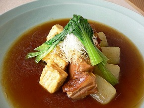 深い緑色の皿に盛られた料理で、中央には煮込まれた角切りの豚肉と揚げ豆腐が並び、その上に青菜と白髪ねぎが彩りよく添えられ、周囲には濃い茶色のだし汁がたっぷりと広がる写真