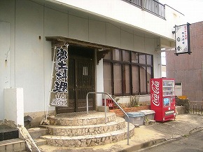 白い外壁の建物に石造りの階段と手すり付きの入口があり、玄関には「郷土料理」と書かれた縦長の暖簾が掛かり、右側には赤い自動販売機と看板が設置された旬香の外観を写した写真