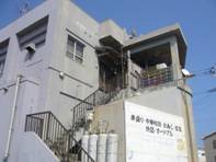 灰色の外壁を持つ二階建ての建物で、外階段が左側にあり、二階のベランダには物干し竿や収納棚が見える店舗外観の写真