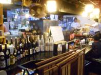 カウンターに多数の酒瓶が並び、奥には調理中の厨房が見える居酒屋風の店内で、暖色の照明が木製のカウンターを照らす写真