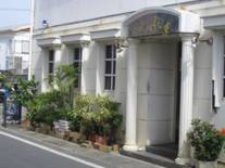 白い外壁に円柱状の装飾柱が並ぶ建物の正面で、入口には半円形の屋根付きドアがあり、周囲に鉢植えの緑が並ぶ「ピュアゴールド 宮花庭」の外観写真