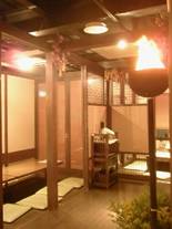 木製の柱と格子で仕切られた和風の個室空間で、畳に座布団が並び、中央に小さなワゴンが置かれ、右手前には炎が灯る装飾的な火鉢がある落ち着いた照明で照らされた店内の写真