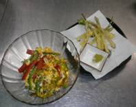 透明なガラス皿に赤と緑の野菜を使った卵とじ風の料理が盛られ、隣には白い紙に載せた細長い野菜の天ぷらと抹茶塩が添えられた和食の写真