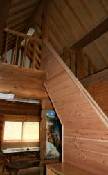 木材で構成された室内に、急な角度の階段が2階の寝室へと続き、天井は三角形の梁が見える吹き抜け構造で、下階には窓と台所を写した写真