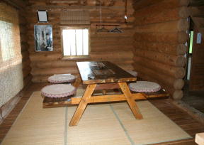 木の丸太を積み重ねた壁に囲まれた室内で、畳敷きの床の中央に木製の長テーブルと両側に座布団が並び、壁には絵画やハンガーが掛けられ、窓から自然光が差し込む落ち着いた雰囲気の居間の写真