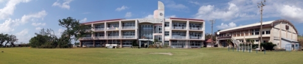 広い芝生のグラウンドを前景に、中央には白い外壁と赤い屋根を持つ三階建ての校舎が建ち、ガラス張りの玄関と時計塔が特徴で、左側には裂けたガジュマルの木、右側には体育館が並び、青空と雲が広がる全景を写した写真