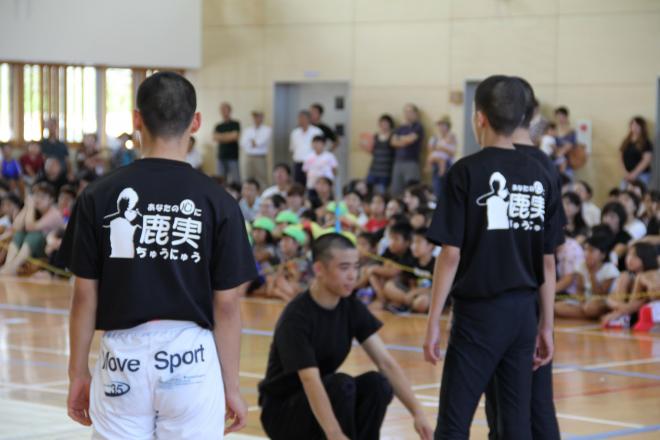 体育館で多くの子どもが見守る中、黒いTシャツを着た少年たちが中央で指導を受けている様子を写した写真