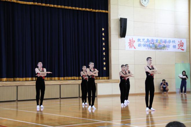 体育館のステージ前で黒と赤の衣装を着た複数の人物が腕を胸の前で交差させて整列している写真。