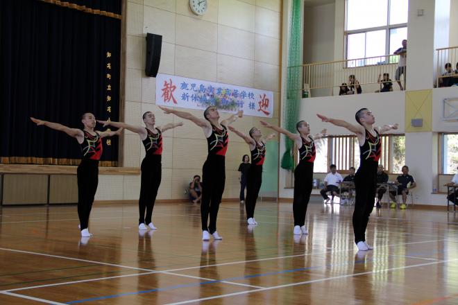 体育館のステージ前で黒と赤の衣装を着た複数の生徒が両腕を左右に大きく広げて立ち、背景には濃紺のカーテンと観客席が見える写真。