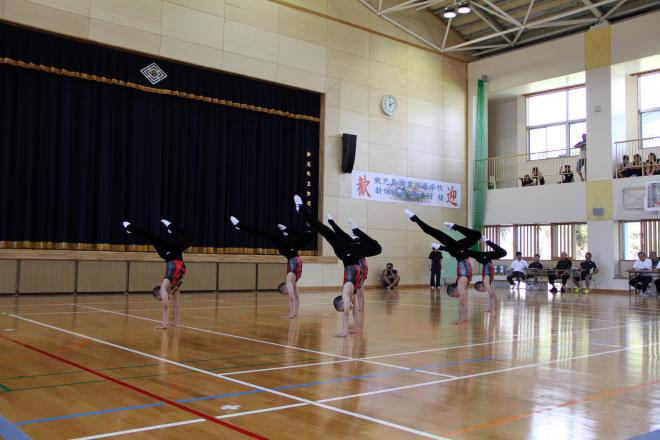 体育館のステージ前で黒と赤の衣装を着た複数の生徒が両腕を伸ばした逆立ち姿勢を揃えて演技しており、背景には濃紺のカーテンと歓迎の横断幕、観客席が見える写真。