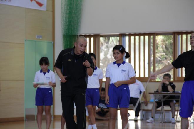 体育館の床の上で、黒い半袖シャツと黒いトレーニングパンツを着用した男性指導者が体操服を着用した女子生徒に指導しており、その後に体操服を着用した2人の生徒が並んでいる様子の写真