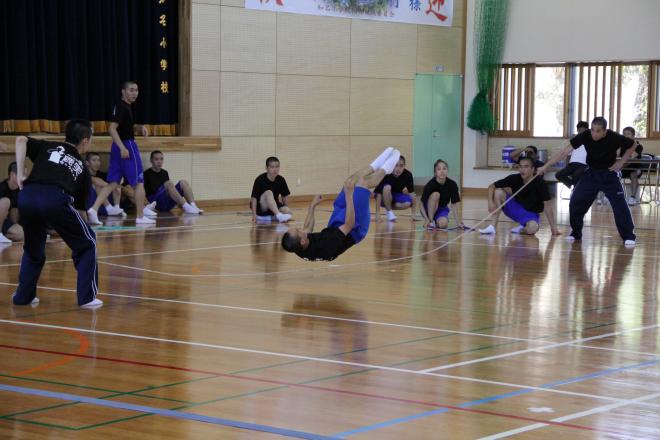 体育館で生徒たちが見守る中、黒いTシャツと青い短パンの男子生徒が縄跳びの下で宙返りのような技を披露している写真