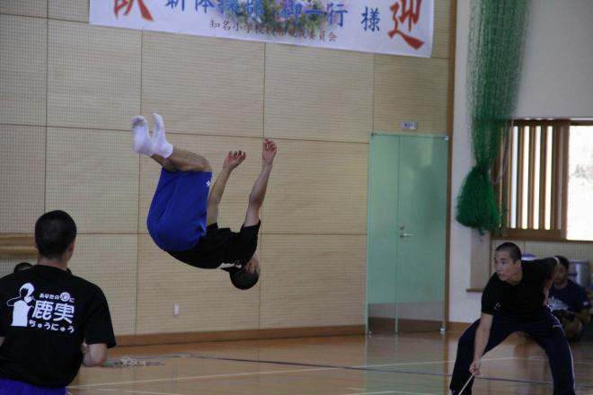 体育館の床で黒いシャツと青い運動ズボンを着た生徒が後方に大きく宙返りを披露している写真