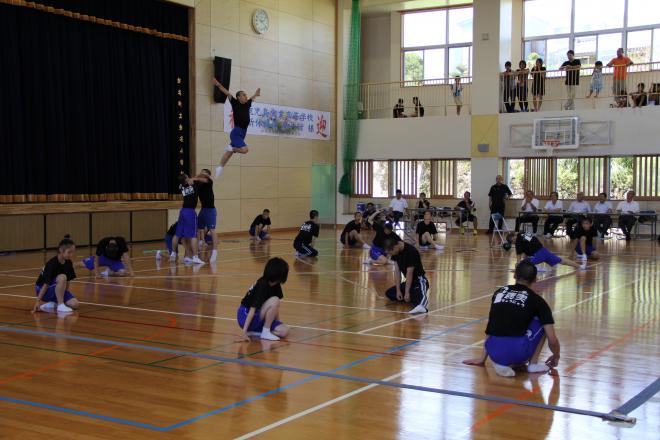 体育館で黒いTシャツと青い短パンの生徒たちが床でポーズを取りつつ、中央では仲間に支えられた生徒が高く持ち上げられて演技を披露している写真