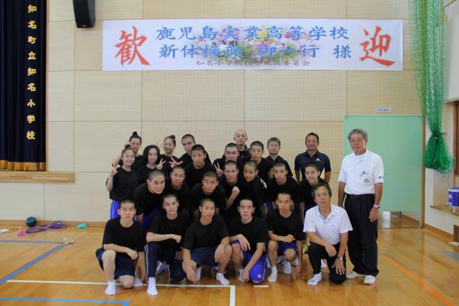体育館のステージ前で黒いシャツと運動着を着た複数の生徒が集合し、後方の壁には「鹿児島実業高等学校 新体操部御一行様 歓迎」と書かれた横断幕の前で記念撮影している写真