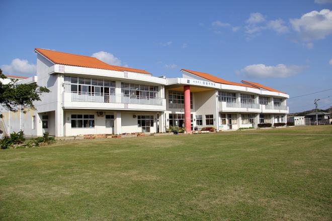 白い二階建ての校舎が広々とした緑の芝生の手前に建ち、鮮やかなオレンジ色の屋根と中央の柱がアクセントとなった、田皆中学校の外観写真