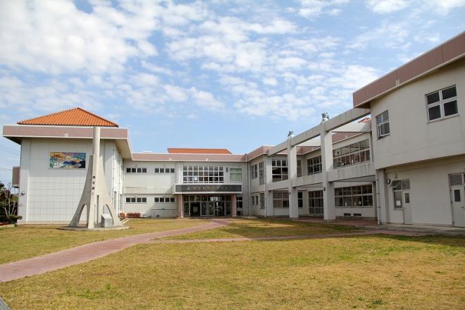 白く明るい外壁を持つ近代的な校舎の建物が、手入れされた芝生の庭と敷地内のレンガ調の小道越しに広がる知名中学校の外観写真