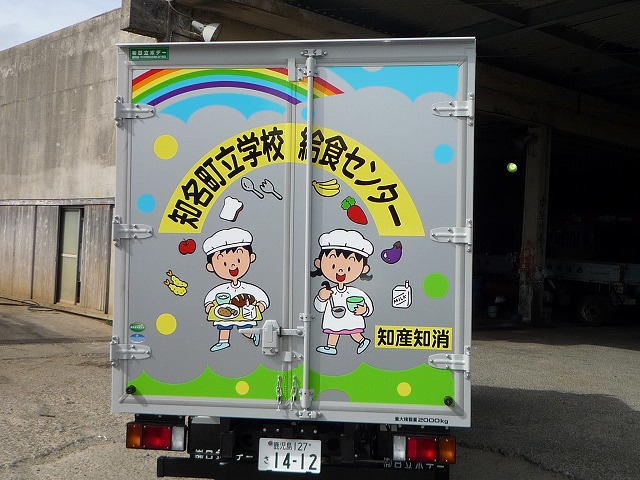 「知名町立学校給食センター」と書かれたアルミ製トラックの荷台の観音扉に、虹と、給食を持って微笑む男の子と、食器を持つ女の子のイラストが描かれ、周囲に食べ物(バナナ、人参、ナス、エビフライ、リンゴ、牛乳パック)の絵や「知産知消」の文字が配された写真