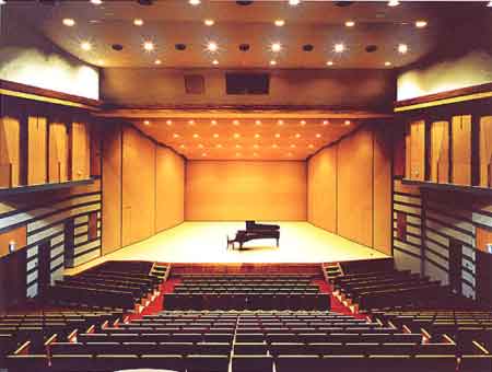 温かみのあるオレンジ色の照明に照らされた、コンサートホールの舞台中央にグランドピアノが一台置かれ、赤絨毯の通路を挟んで黒い座席が段状に並ぶ客席を上から見下ろす写真