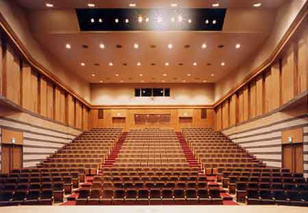 明るい木目調の壁と天井を持ち、赤色の絨毯が敷かれた中央通路を挟んで茶色の座席が左右対称に並び、後方に向かって段状に傾斜しているホールの客席を舞台上から捉えた写真