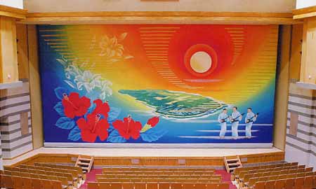 コンサートホールの舞台正面で、壁一面に原色を多用したハイビスカスと白いユリなどの花々、太陽と海、そして三味線を持ち白い衣装を着た3人の人物が描かれたホールの緞帳の写真
