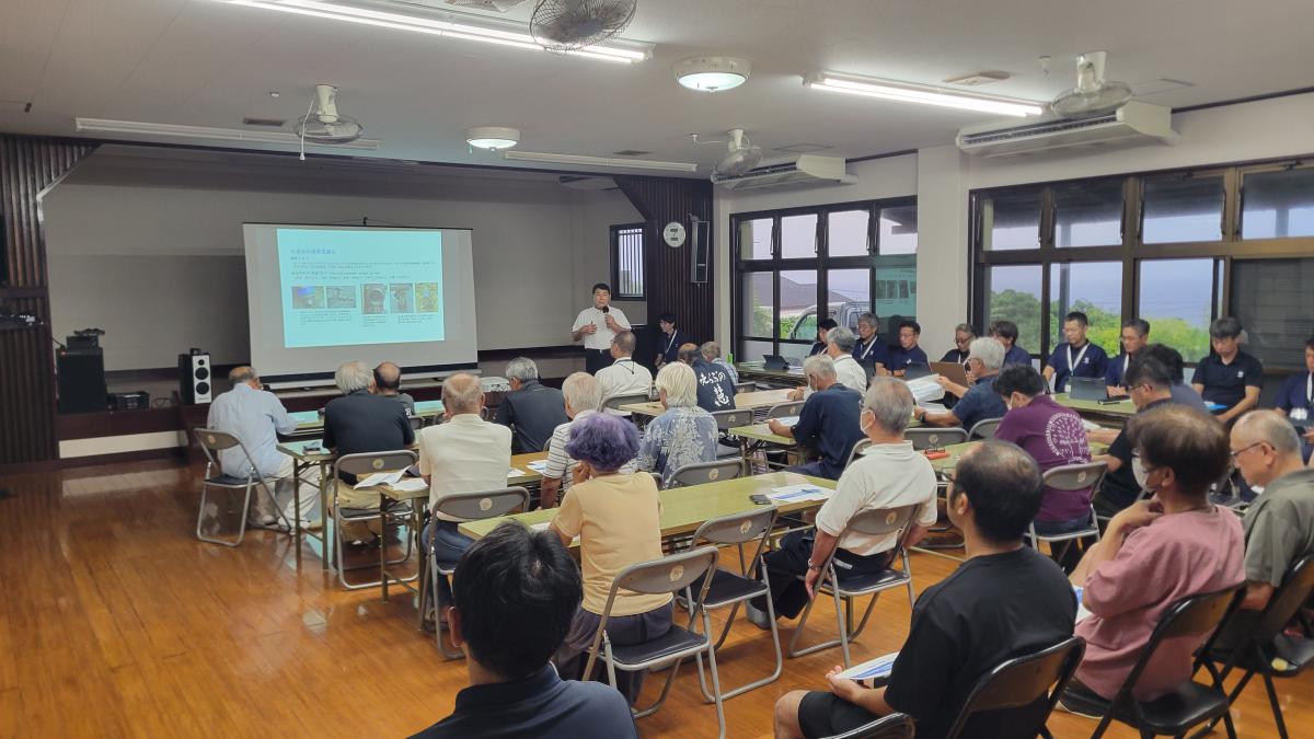 会議室で複数の人物が長机に着席し、前方のスクリーンに資料が投影され、発表者がマイクを持って説明している様子の写真