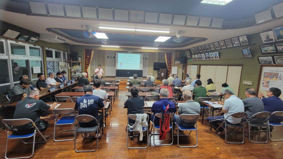 会議室で複数の人物が長机に着席し、前方のスクリーンに資料が投影され、発表者が説明を行っている様子を後方から写した写真