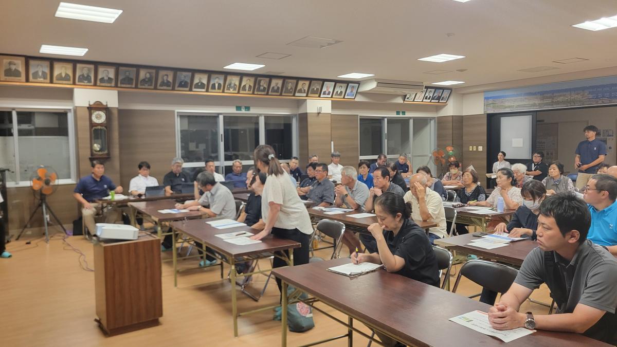 会議室で複数の人物が長机に着席し、資料を広げて説明を聞いている中で、参加者の女性が立ち話をしている写真