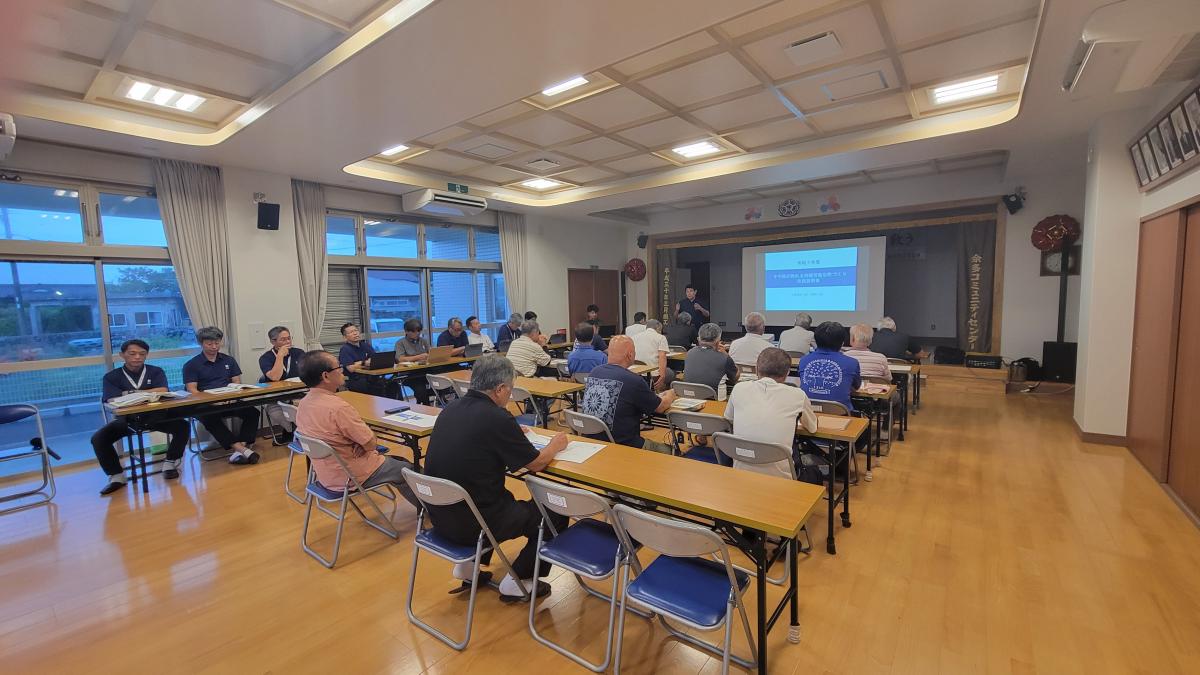 会議室に長机と椅子が配置され、中央で一人の男性がプロジェクターのスクリーンに向かって多くの参加者に説明を行っている様子の写真