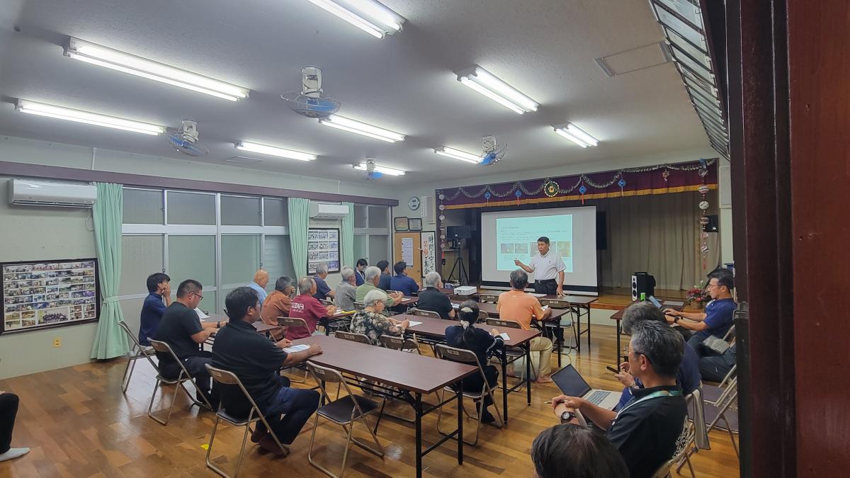 明るい会議室で、一人の男性がプロジェクターのスクリーンを背にして参加者のグループに向かい立って話し、参加者の方々は机と椅子に座って熱心に耳を傾けている様子の写真