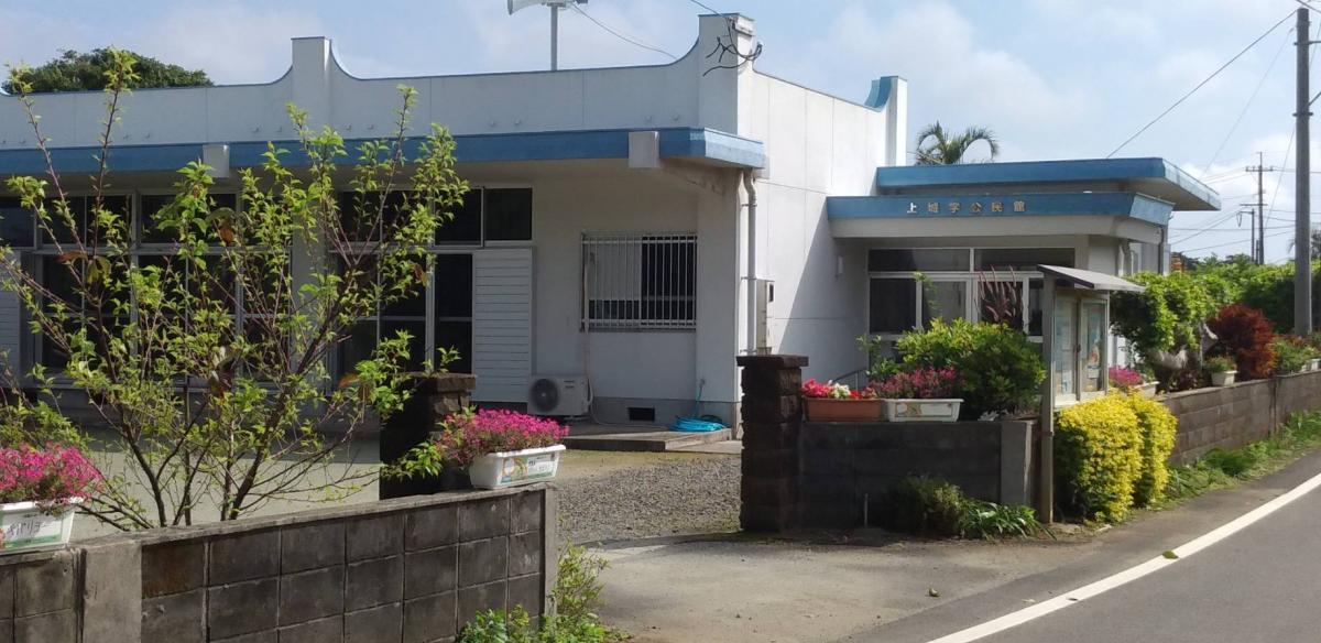 白い外壁に青い縁取りが施された平屋の建物が道路沿いに建ち、右側にはガラス張りの掲示板が設置された地域施設の写真。