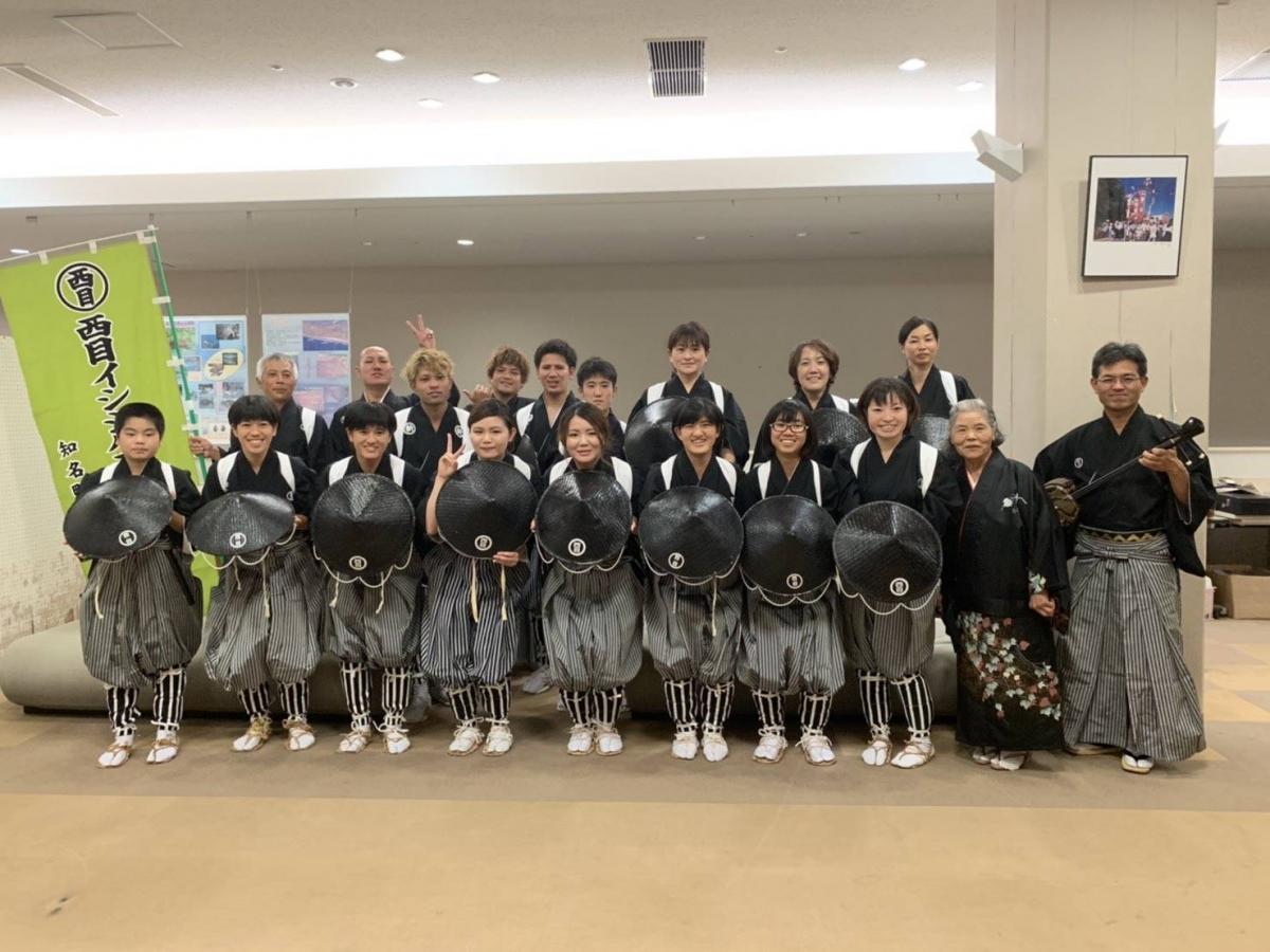 黒と白の縞模様の袴に黒い羽織を着用し、胸に丸い飾りを付けた一団が室内で整列しており、左側には西目イシシハカマ踊の旗が見える集合写真。