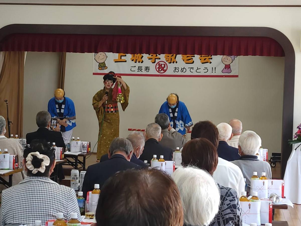 舞台の上で黄色の和装を着た人物が飾りを持って立ち、後方には青い法被姿の二人が並び、前方には長寿祝いの横断幕が掲げられた室内で多くの参加者が着席している敬老会の写真。