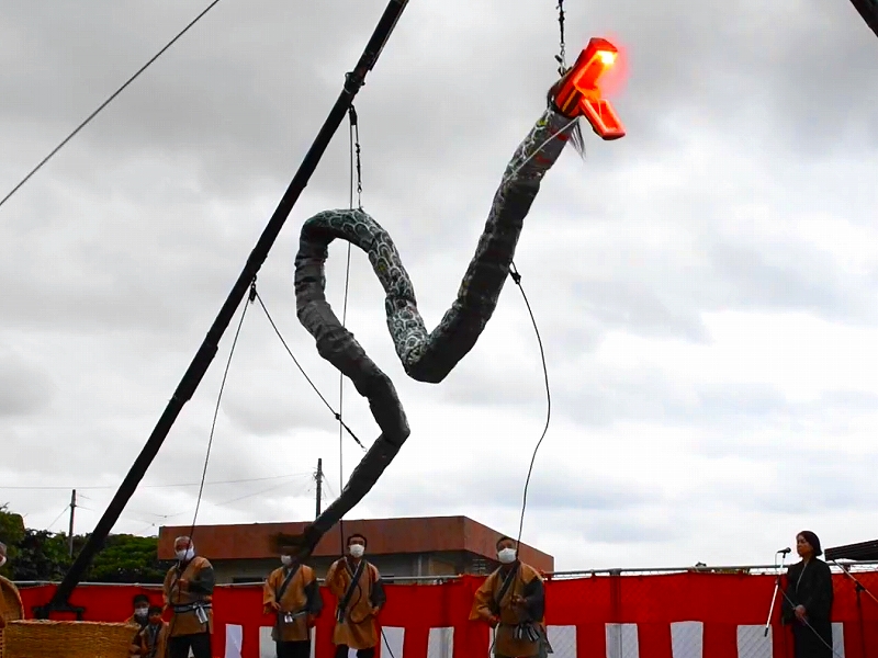 赤い光を放つ大蛇の巨大な模型が空中に吊り下げられ、伝統衣装を着た複数の人物がその下で見守る屋外祭礼の様子を捉えた写真。