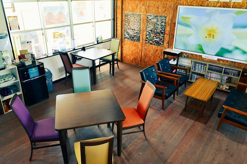 木目の床が広がる室内に、カラフルな椅子を備えたテーブルが複数配置され、奥にはソファと木製のローテーブルが並び、壁には大きな花の写真と本棚が設置されたくつろぎスペースの写真。