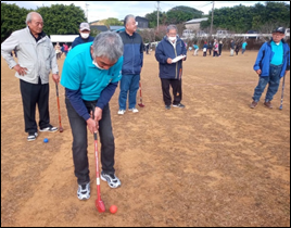 青いシャツを着た人物が赤いスティックで赤いボールを打とうとしており、周囲には同様のスティックを持った数人が立って見守っている写真。