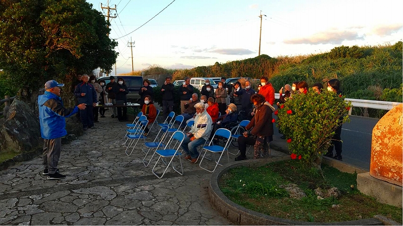屋外の石畳の広場で、青いジャンパーを着た人物が前方で話すのを、折りたたみ椅子に座っている人々と立っている人々が聞いている写真。