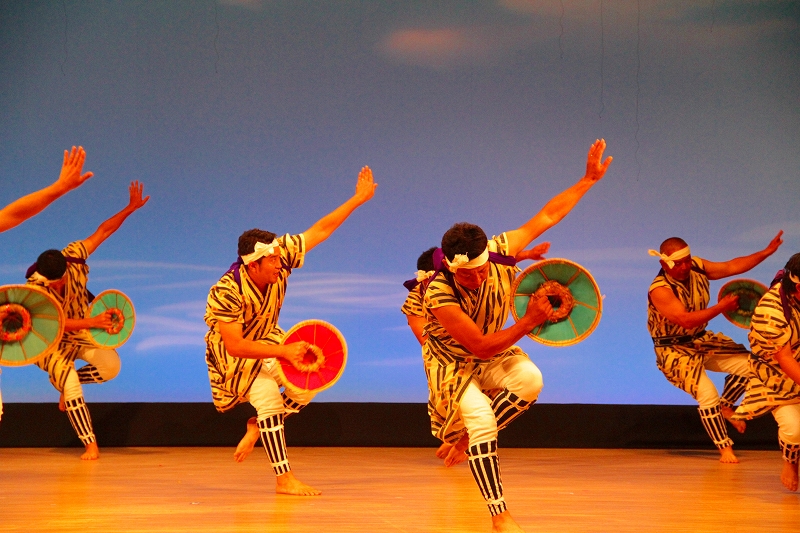 舞台上で縞模様の衣装を着た複数の踊り手が、片腕を高く上げ片腕を前に伸ばしながら、赤や緑の円形の道具を手に持って力強いポーズを取っている伝統舞踊の様子を捉えた写真。