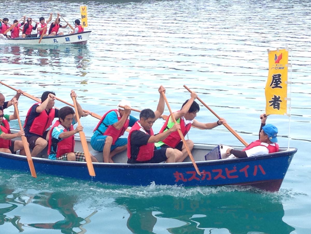青い船体に赤い文字が描かれたボートに複数の人々が赤いライフジャケットを着けて乗り込み、オールを力強く漕いで進む様子が水面に映り、右側には黄色い旗が立ち、奥には同様のボートが見えるくり舟競争の写真。
