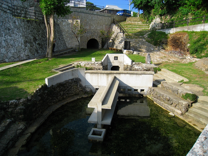 石造りの斜面に囲まれた緑の庭園内に、コンクリートで整備された湧水池があり、中央には渡し板状の構造物が設置され、奥にはトンネル状の水源口と石碑が見え、周囲には階段や遊歩道が整備された水場を写した写真