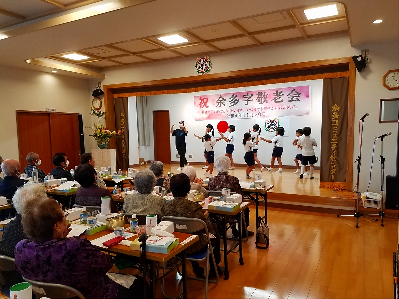 室内の舞台で「祝 余多字敬老会」と書かれた横断幕が掲げられ、前方では子どもたちが並んで踊りを披露し、手前のテーブルには食事や飲み物が並び、複数の高齢者が座って鑑賞している写真。