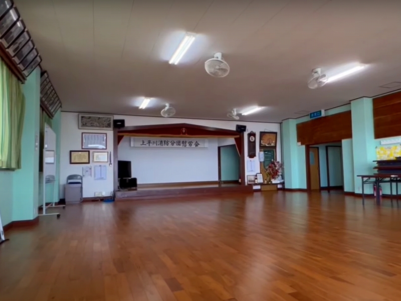 木目の床が広がる広い室内に小さな舞台が設けられ、壁には額縁や掲示物が並び、天井には蛍光灯と扇風機が取り付けられた地域集会所の写真。