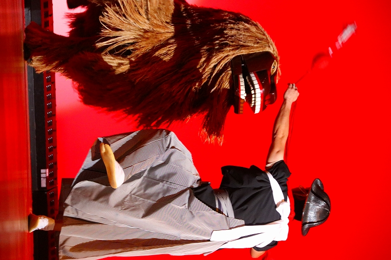 赤い舞台背景の中、和装の演者が獅子舞の大きな獅子頭に向かって棒を振り上げ、躍動感ある伝統芸能の一場面を写した写真