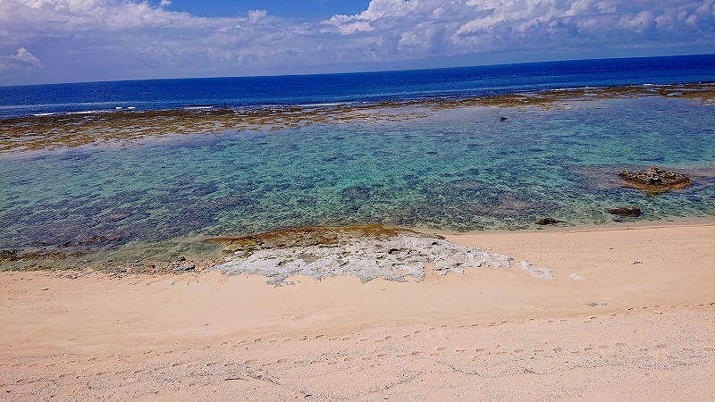 白い砂浜の先に透明度の高い浅瀬が広がり、海底の岩やサンゴが透けて見え、奥には濃い青の海と雲が浮かぶ青空が続く静かな海岸風景の写真。