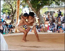 屋外の土俵で二人の子どもがまわしを締めて組み合い、周囲には観客が座って見守っている相撲大会の様子を捉えた写真。