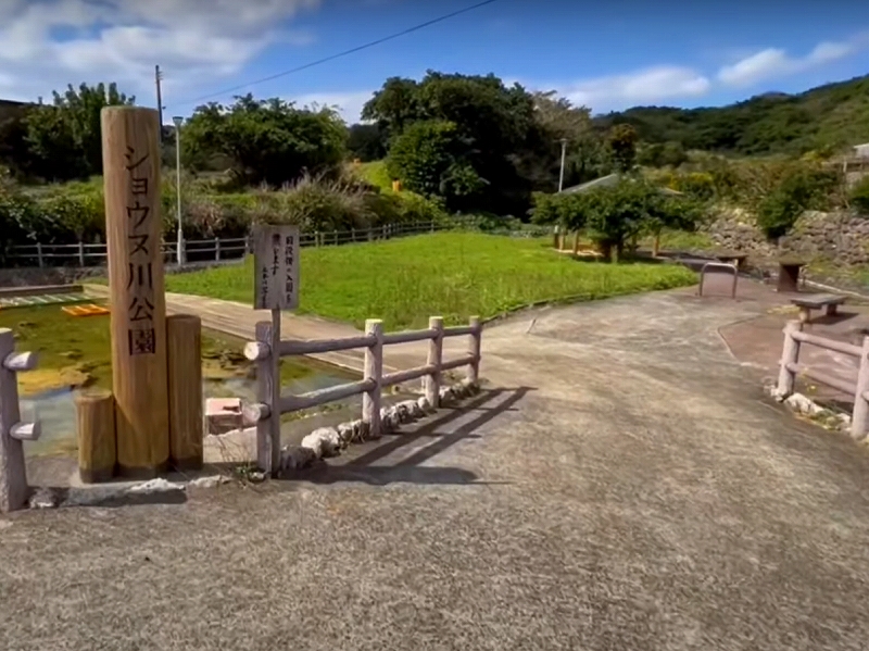 木製の案内柱に「ショウス川公園」と刻まれた文字が立ち、周囲には柵で囲まれた小川と緑の芝生が広がり、奥には木々とベンチが配置された自然豊かな公園の写真。
