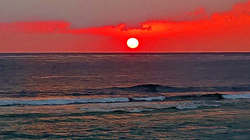 赤く染まる雲の下で真円の夕日が水平線近くに沈み、穏やかな波が広がる海面に鮮やかな赤と橙の光が映える風景の写真。